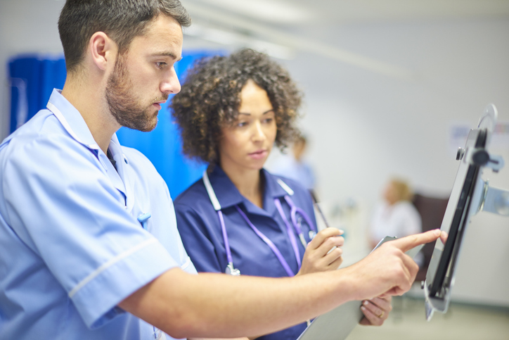 A male nurse looks at a computer screen with a female nurse watching on