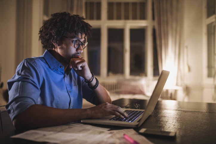 A man is sat looking at his laptop in the dark with the screen glowing