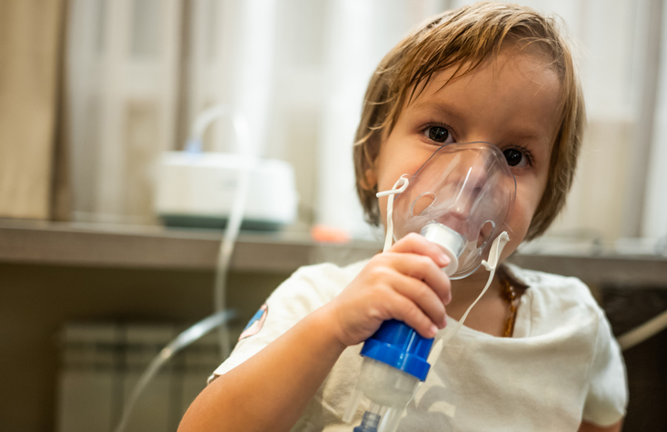 A young boy sits in a hospital gown with a breathing device on